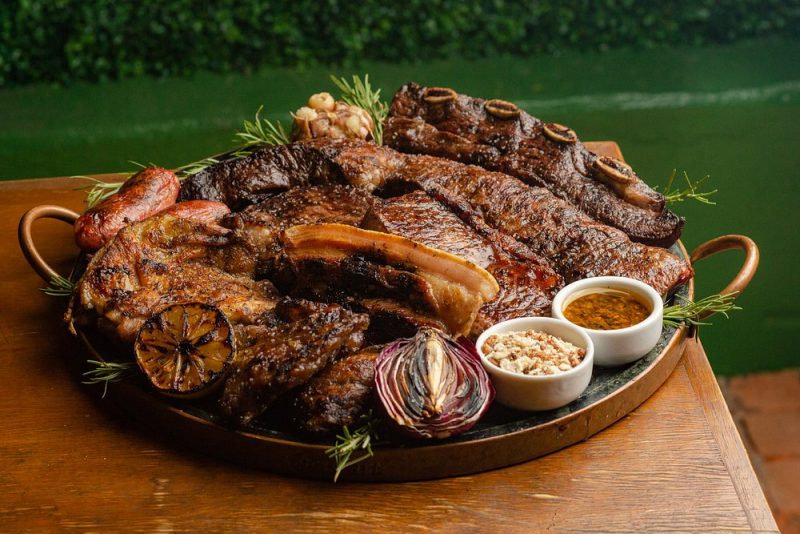 Foto de uma bandeja com grandes carnes e pequenos potes de molho sobre uma mesa de madeira, no restaurante Los Troncos. 