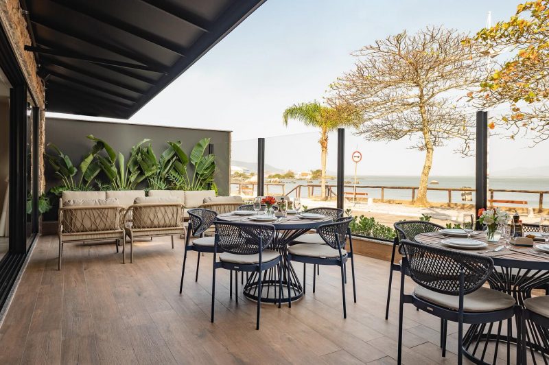 Foto do deck externo do restaurante Rancho Açoriano, com chão de madeira, poltronas de vime e mesas redondas. O restaurante é uma das opções de onde comer em Coqueiros e tem vista para o mar. 