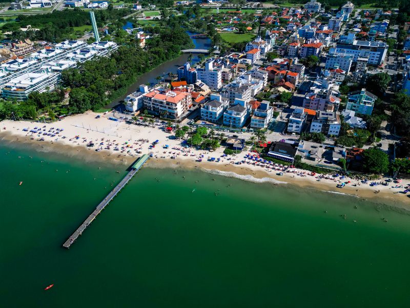 Foto aérea da Praia de Canasvieiras. O mar é esverdeado e no canto esquerdo há uma passarela de madeira que parte da areia da praia e segue pelo mar. A areia da praia é clara e está cheia de guarda-sóis. Atrás dela, há prédio, um córrego do lado esquerdo e vegetação. 