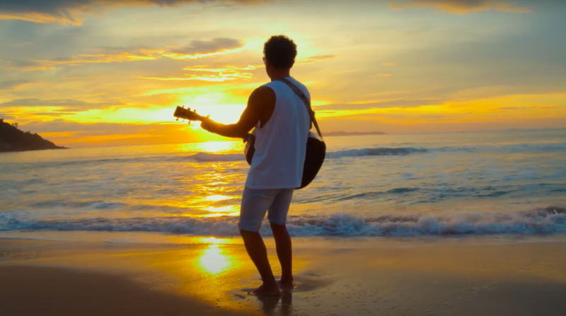 Frame de videoclipe mostra cantor Nego Joe de costas, tocando violão e olhando o mar