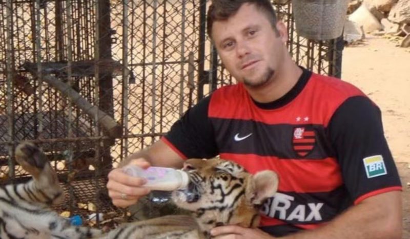 Foto mostra Sérgio Bala com uma camiseta do Flamengo enquanto ele dá mamadeira para uma onça. Ele é um homem branco, de cabelos castanhos e cavanhaque