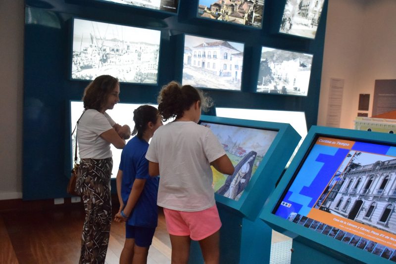 Foto de duas crianças e uma mulher adulta olhando a tela de um totem interativo do Museu de Florianópolis. Ao fundo, há várias outras telas com fotos de diferentes épocas da cidade. 