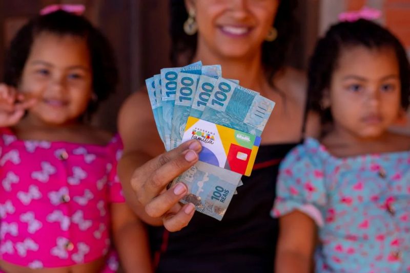 Mulher com suas filhas segurando o cartão do bolsa família e um leque de dinheiro 