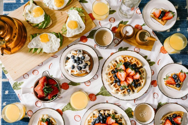 Mesa de café da manhã decorada e repleta de pratos deliciosos, como torrada com ovos, waffle com frutas, tigela de salada de frutas, sucos e café 