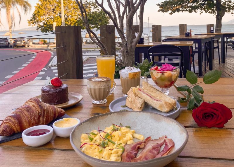 Foto de uma mesa de madeira, com vários pratos em cima. Em um dos pratos há ovos mexidos e bacon. Ao lado, um croissant e um pequeno bolo de chocolate. Em outro prato, um sanduíche. Sobre a mesa há também uma taça com frutas, xícaras de cafés, copo de suco de laranja e uma rosa. Ao fundo, aparece parte da via, com árvores e o mar atrás. 