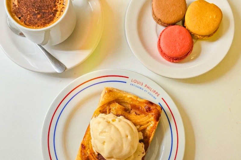 Foto de uma mesa branca com dois pratos e uma xícara de chocolate. Os dois pratos têm doces. Em um deles está escrito o nome da cafeteria. 