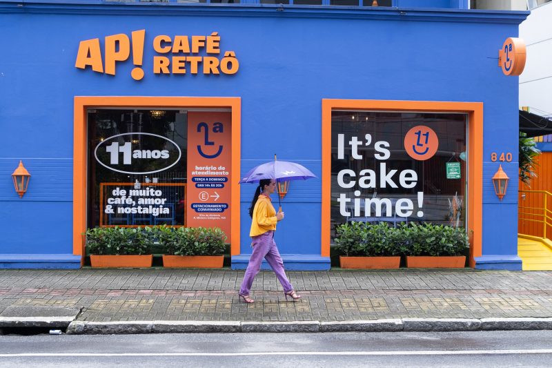 Mulher jovem usando guarda-chuva passando em frente a um dos cafés em Joinville, com fachada azul e o nome AP! Café Retrô escrito em laranja. 