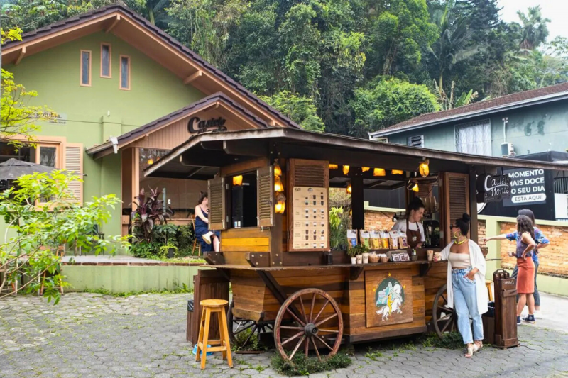 Carruagem de madeira, com janelas e um balcão onde estão expostos os cafés. Uma mulher jovem está apoiada nela, segura um copo de café e olha para a atendente. Ao fundo, um quintal e uma casa verde onde estão três pessoas. 