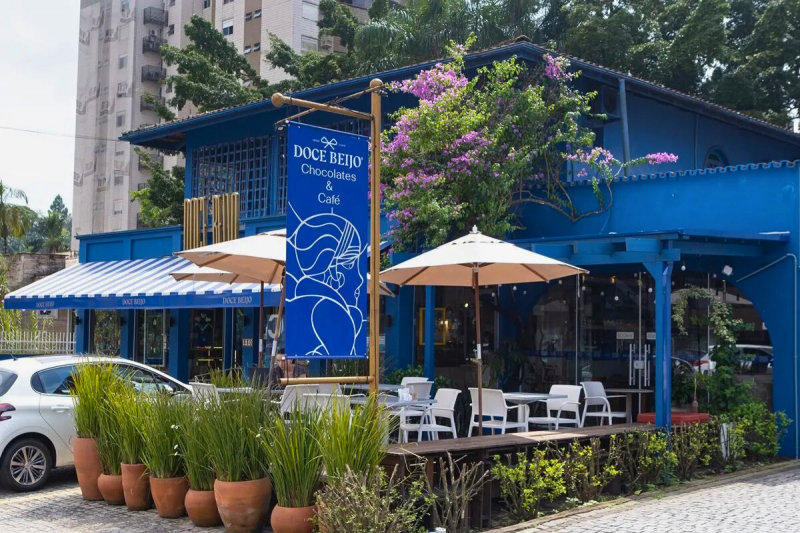 Sobrado azul da chocolateria em Joinville. Na frente, uma área de varanda com mesas e cadeiras brancas e ombrelones. À frente, alguns vasos com folhagens. 