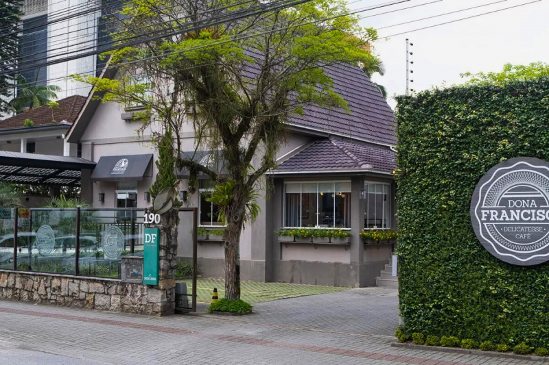 Fachada de um dos cafés em Joinville. A casa é cinza, com janelas de vidro e floreiras. À frente da casa, uma árvore. Parte do muro é repleto de plantas e tem a logomarca da Dona Francisca Delicatesse Café. 