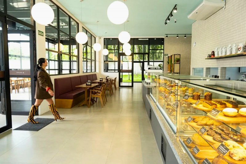 Mulher jovem entrando em uma cafeteria ampla, com tons claros e uma bancada cheia de quitutes. 