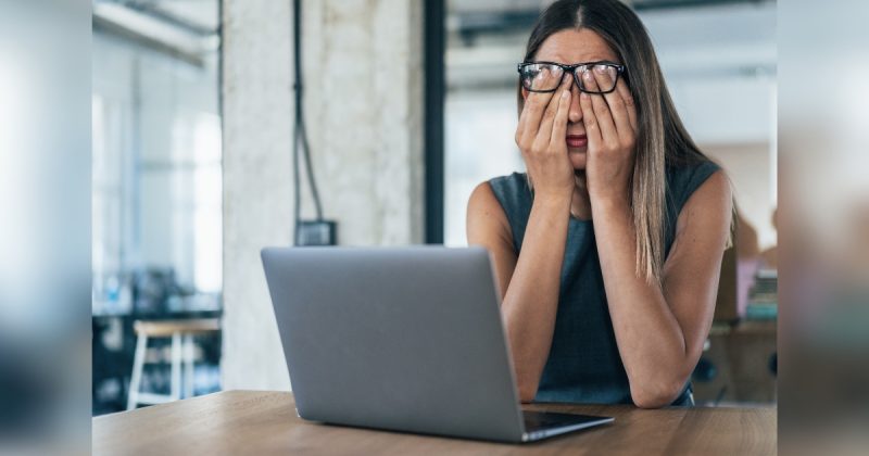 Mulher com óculos no rosto e de frente a computador com mãos no rosto