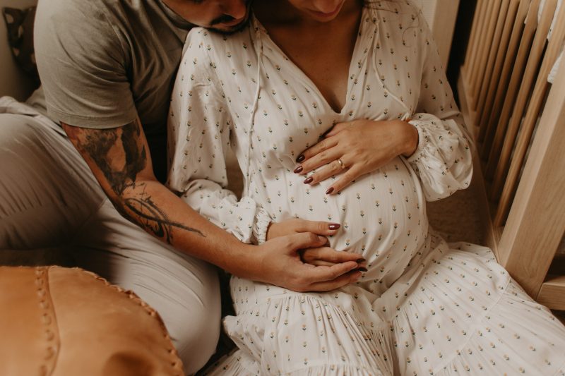 Imagem de casal, com foco na mulher grávida que está com suas mãos e do companheiro na barriga