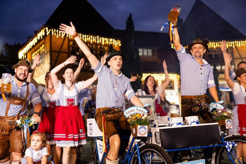 Foto do desfile na rua da Festa Pomerano. Homens e mulheres vestidos com trajes típicos estão com as mãos erguidas, seguram grandes canecos de chope sorriem. Atrás o pórtico de entrada de Pomerode, com luzes de Natal. 