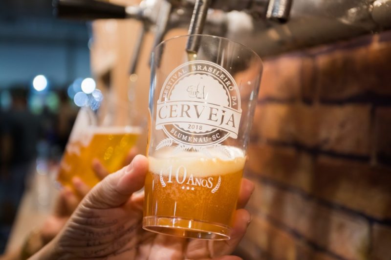 Foto da mão de um homem, de pele clara, segurando um copo de vidro abaixo de uma torneira de chope. No copo, escrito em branco: Festival Brasileiro da Cerveja 2018 - Blumenau -SC - 10 anos. 