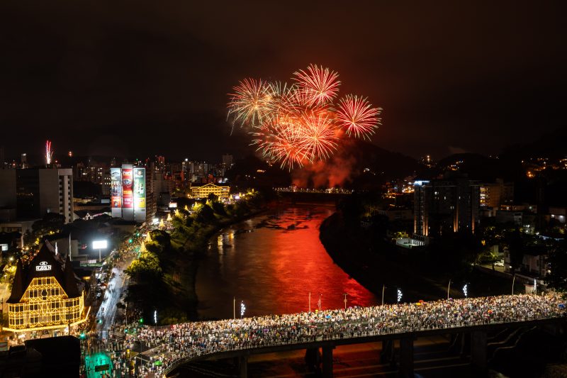 Foto da queima de fogos em Blumenau. Milhares de pessoas estão sobre a Ponte de Ferro, que passa por cima do Rio Itajaí Açú. Ao fundo, fogos em formato circular e vermelho no céu. Nas laterais do rio, alguns prédios iluminados. 