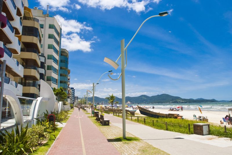 Imagem de um calçadão com postes de luz, faixa de areia com a praia ao fundo