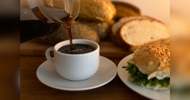 Xícara de café com mais café sendo colocado e um sanduíche do lado