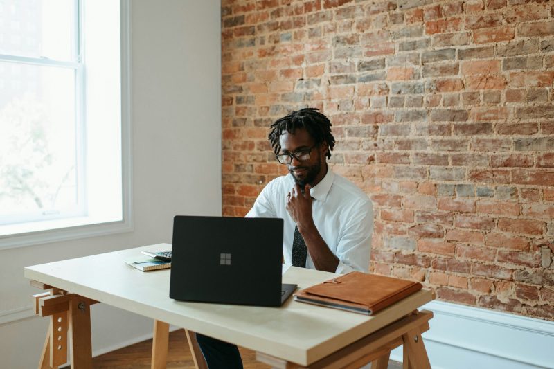 Homem negro sentado em frente a um notebook, com a mão no queixo, em uma sala bem iluminada 