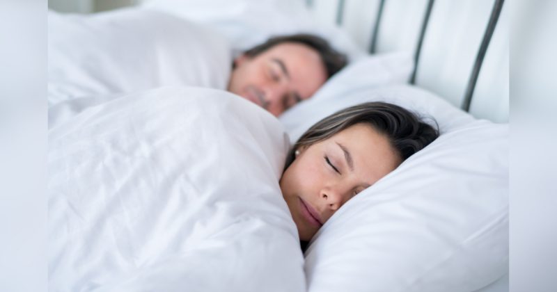 Duas pessoas dormindo em cama com lençóis na altura do pescoço