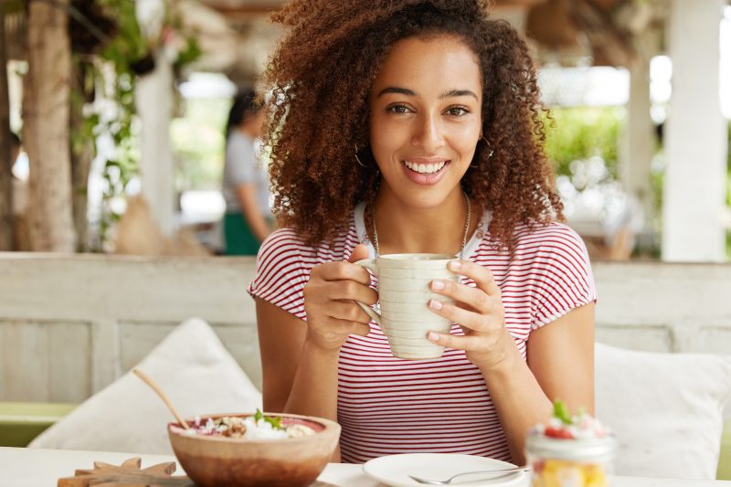 Mulher negra segurando uma xícara de café em frente de uma mesa do café da manhã 