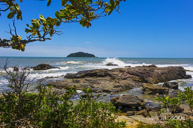 Visto da praia a partir da vegetação local, com árvores, pedras antes da areia e rochas no mar 