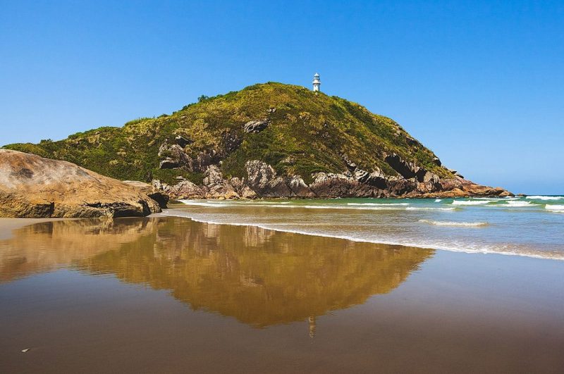 Imagem de uma praia com um morro coberto de vegetação natural ao fundo, com um farol visto ao longe