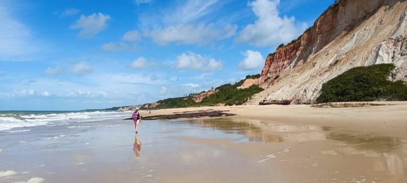 Mulher caminhando em praia cercada por grandes falésias coloridas 