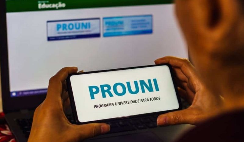 Para ilustrar o Prouni 2025, um homem segura o celular e, na tela, aparece a mensagem "Prouni (Programa Universidade Para Todos)