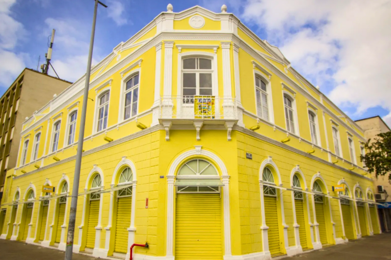 Casarão amarelo de dois andares e de esquina no centro de Joinville. Os ornamentos em torno das janelas e portas são na cor branca. 