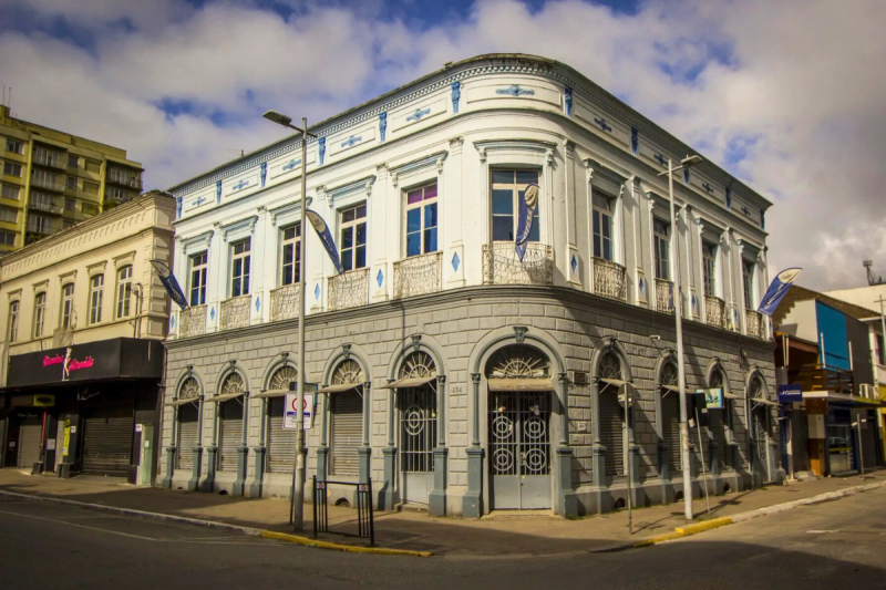 Prédio de esquina e de dois andares. A parte superior é azul claro com detalhes em azul escura. Há 10 janelas com ornamentos sobre elas e pequenas varandas. A parte superior é cinza. Sobre cada uma das 10 portas há um semiarco decorativo. 