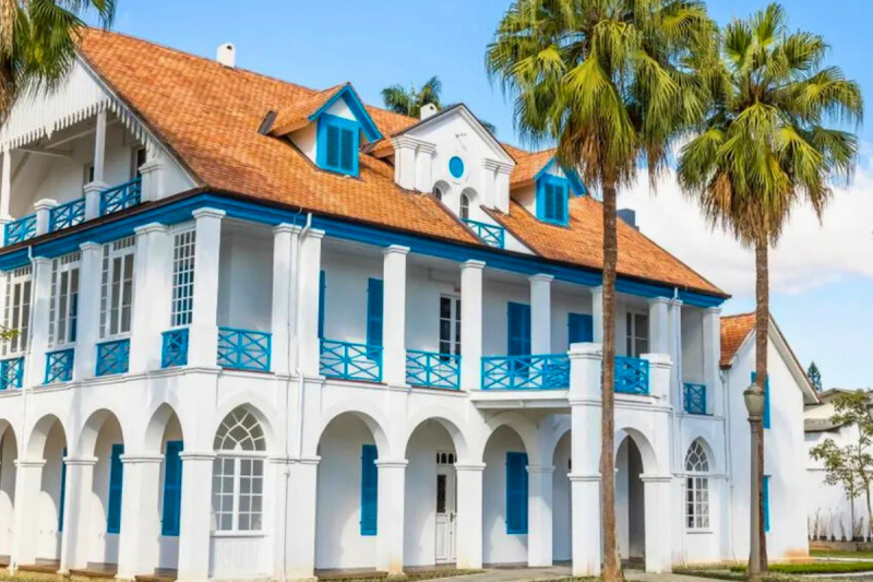 Casarão de três andares, branco e com 10 colunas. As janelas e as madeiras do parapeito da sacada que circunda o edifício são azuis. A frente da casa há duas palmeiras. 