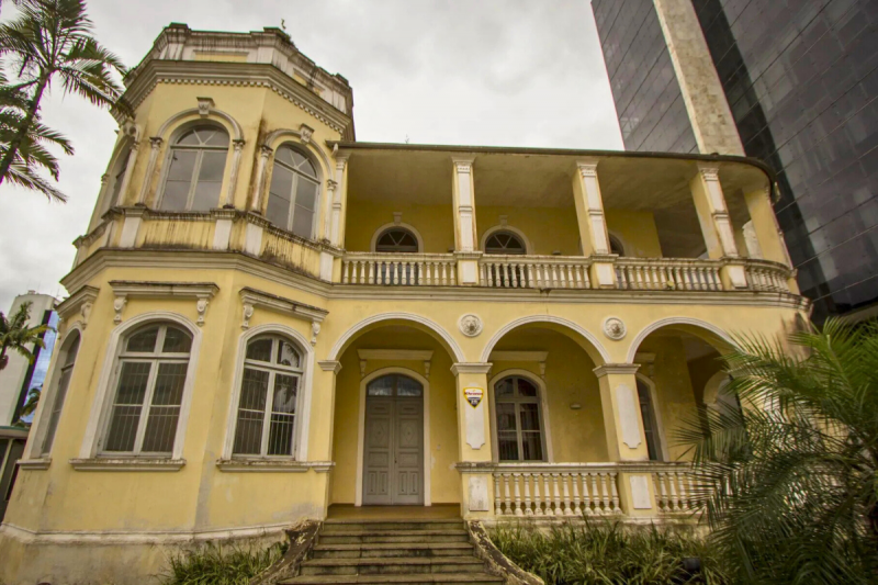 Sobrado bege com balaústres e uma torre larga do lado esquerdo com ornamentos. Do lado direito da foto é possível ver parte do prédio do banco do Brasil todo de vidro e em estilo moderno. 