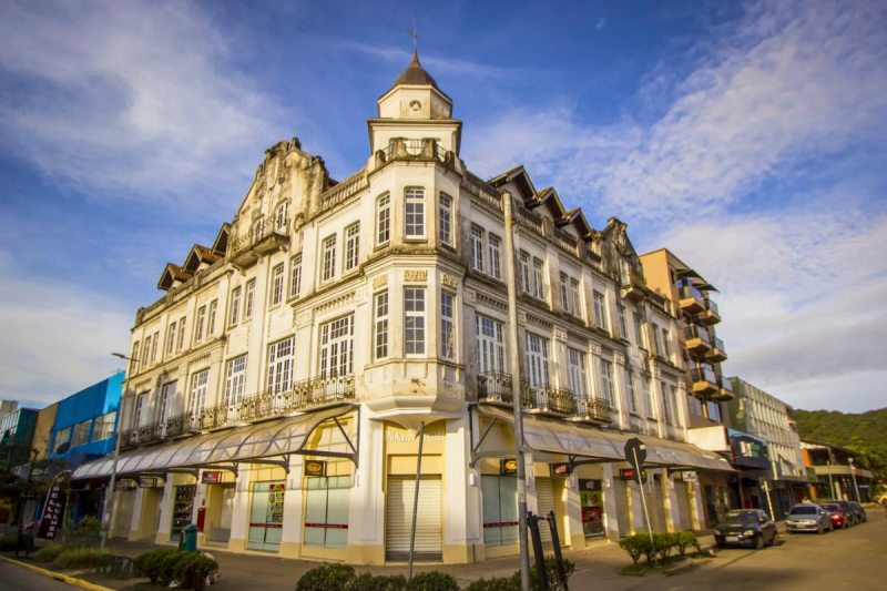 Prédio de três andares, de esquina, com mais de 30 janelas e uma torre no centro. O prédio possui muitos ornamentos e na parte debaixo funcionam lojas de comércio. 