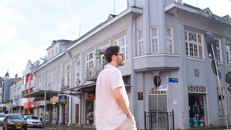 Homem jovem de pele branca com fones de ouvido em frente a um prédio antigo cinza na Rua do Príncipe, no Centro de Joinville. 