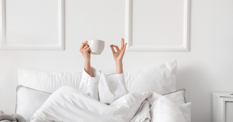 Pessoa segurando café com mão e na outra fazendo simbolo debaixo das cobertas