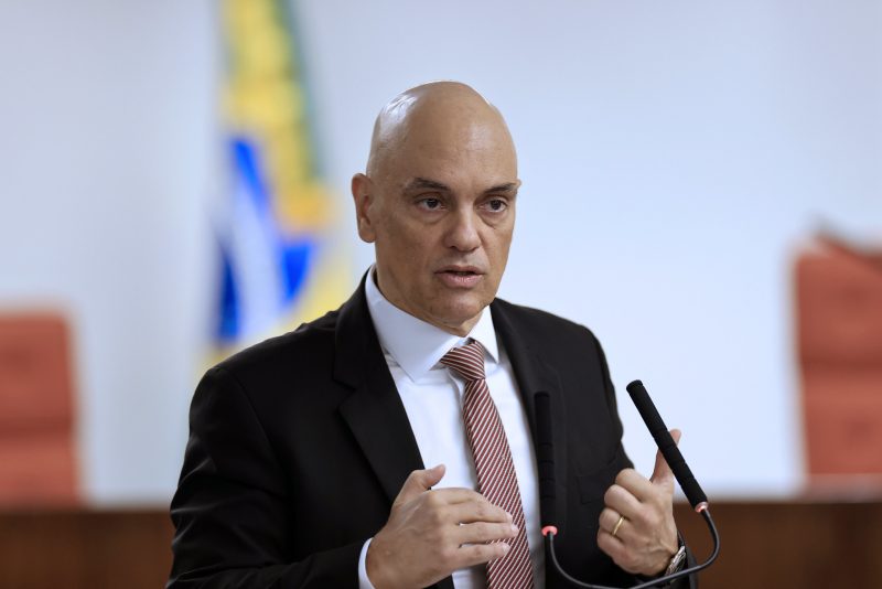 Alexandre de Moraes, homem branco e de terno em frente a um microfone e uma palestra. Cadeiras e a bandeira do Brasil aparecem desfocadas no fundo