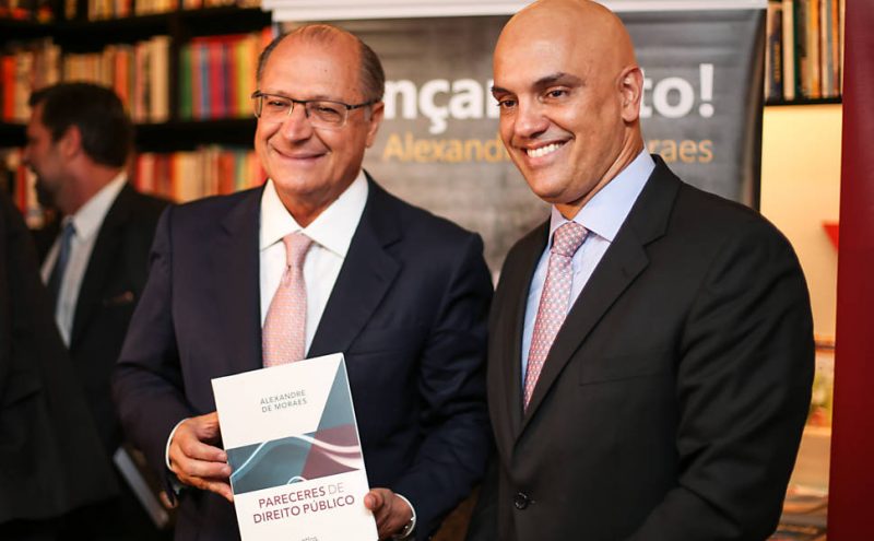 Dois homens brancos usando terno em uma livraria. Na esquerda, Geraldo Alckmin, segura o livro de Alexandre de Moraes, que está à direita da foto