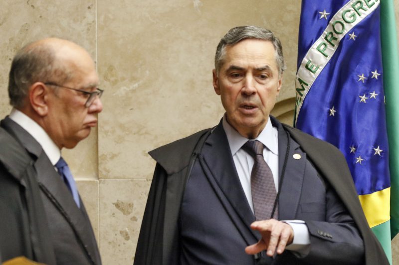 Foto de dois homens brancos, com torga do STF. Na esquerda, de perfil está Gilmar Mendes e ao centro está Luis Roberto Barroso, em frente a bandeira do Brasil 