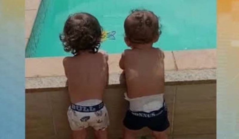 Foto mostra bebês gêmeos de costas apoiados na beira da piscina