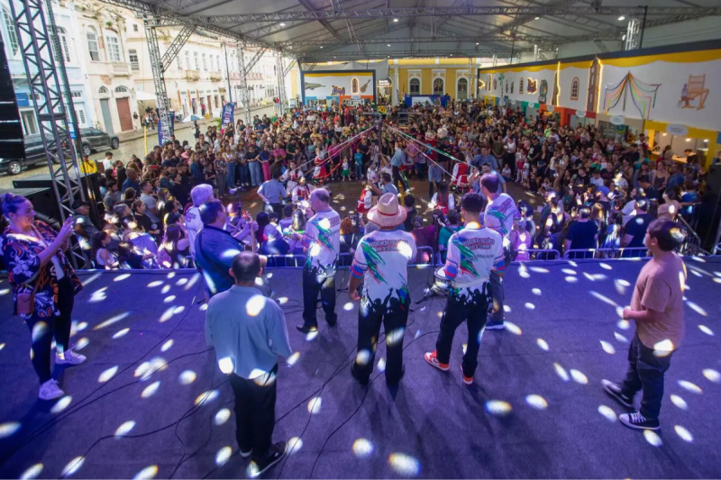 Em primeiro plano, um palco onde estão nove homens e uma mulher. Eles estão de costas para a foto. À frente deles uma plateia com centenas de pessoas. Próximo ao palco, algumas pessoas dançam entrelaçando fitas coloridas presas a um pau de madeira. O espaço é coberto. 