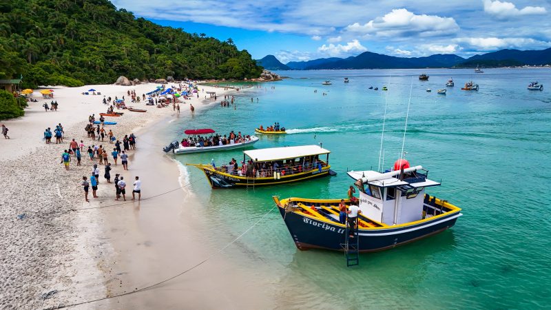 Foto de dois barcos de tamanho médio e uma lancha ancorados na areia branca da Ilha do Campeche, em Florianópolis (SC). No lado direito o mar de água cristalina esverdeada. No canto esquerdo, a vegetação cercando a areia, por onde andam algumas pessoas. 