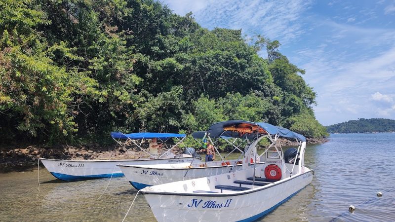 Foto de três barcos brancos parcialmente cobertos com um toldo azul. Na lateral do barco escrito: JV Ilhas. Eles estão ancorados em águas tranquilas da Ilha do Pelado, em Paraty (RJ). No canto esquerdo, bastante vegetação.