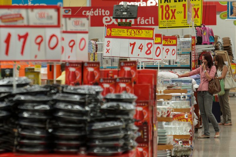 Imagem de um supermercado com placas de preço e pessoas com carrinhos fazendo compras 