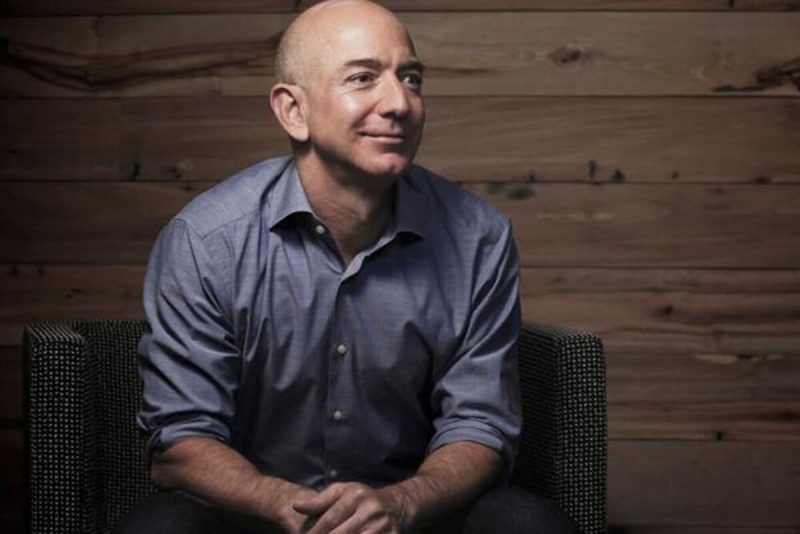 Jeff Bezos, homem branco careca, sentado em frente a fundo neutro, com as mãos apoiadas nas pernas 