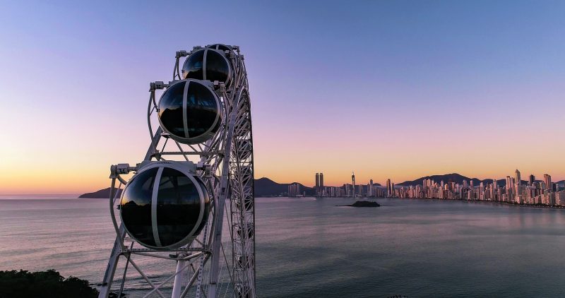 Foto externa de algumas das cabines da Roda Gigante de Balneário Camboriú, com vidro preto e fixadas em um grande aramado de metal. Ao fundo, o extenso mar e os altos prédios da orla da Praia Central. O céu está em cores de azul e laranja, ao entardecer. 