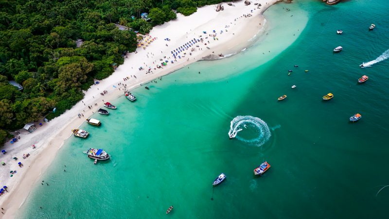 Foto aérea da Ilha do Campeche. Na maior parte da foto, aparece o mar de águas claras esverdeadas, onde estão vários barcos coloridos que transportam as pessoas até a ilha. Cercando o mar, uma extensa faixa de areia branca, com dezenas de guarda-sóis coloridos. Atrás da areia, vegetação. 