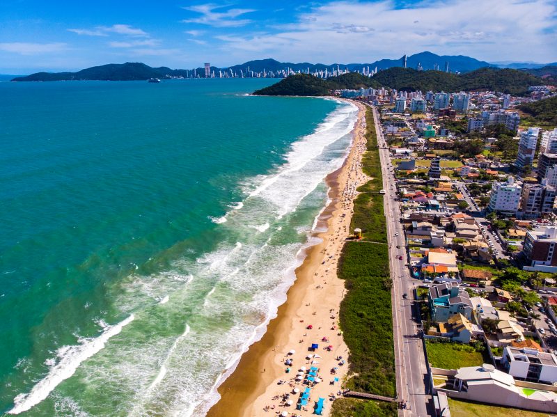 Foto aérea da Praia Brava, em Itajaí. Do lado esquerdo da foto, o imenso mar de cor azul esverdeada com ondas. Do lado direito, uma extensa faixa de areia bege com vários guarda-sóis coloridos. Cercado a areia, uma faixa de vegetação de restinga. Após a faixa, uma avenida asfaltada e muitas casas e prédios. Ao fundo morros verdes e a vista de longe dos altos prédios de Balneário Camboriú. 