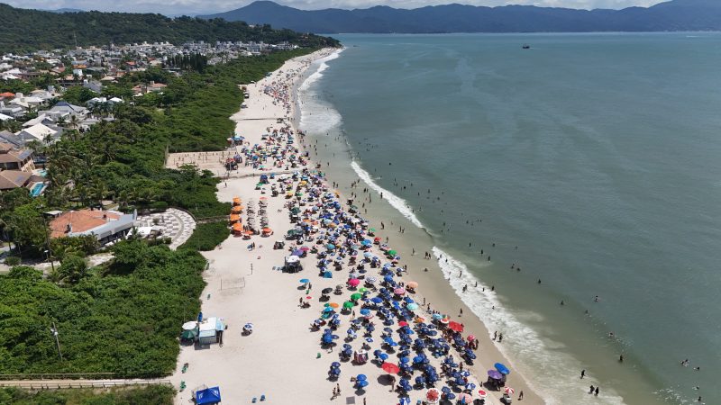 Foto aérea de Jurerê Internacional. Do lado direito, o mar azul com poucas ondas e dezenas de pessoas na água. Do lado esquerdo, a areia branca com centenas de guarda-sóis coloridos, cercada por uma extensa e larga faixa de vegetação de restinga. Atrás delas, muitas construções. No horizonte, morros verdes.