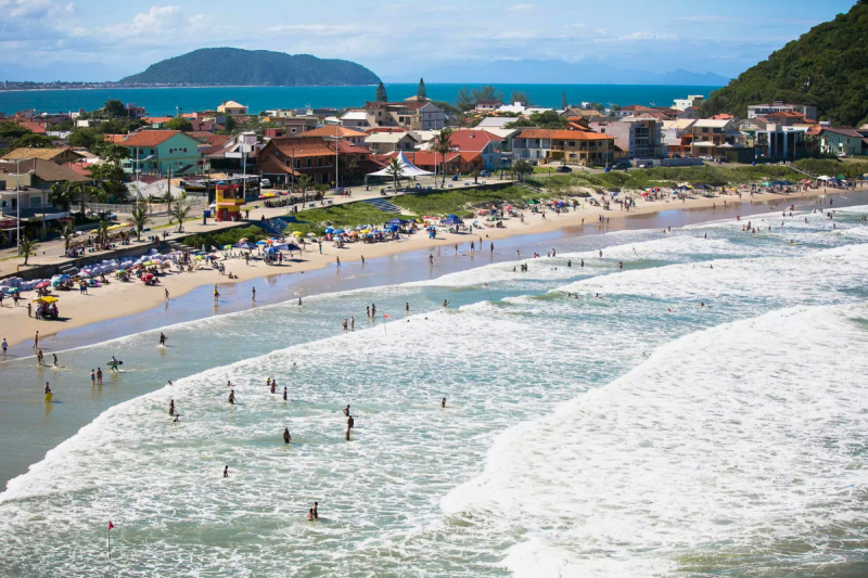 Foto aérea da Prainha, em São Francisco do Sul. Pegando quase toda a foto, o mar de águas claras e ondas, onde estão dezenas de pessoas. Cercando ele, a areia clara onde estão vários guarda-sóis coloridos. Em torno da areia, uma faixa de areia de restinga. Atrás deles, uma rua com casas e mais ao fundo, o mar de outra praia e um morro verde. 